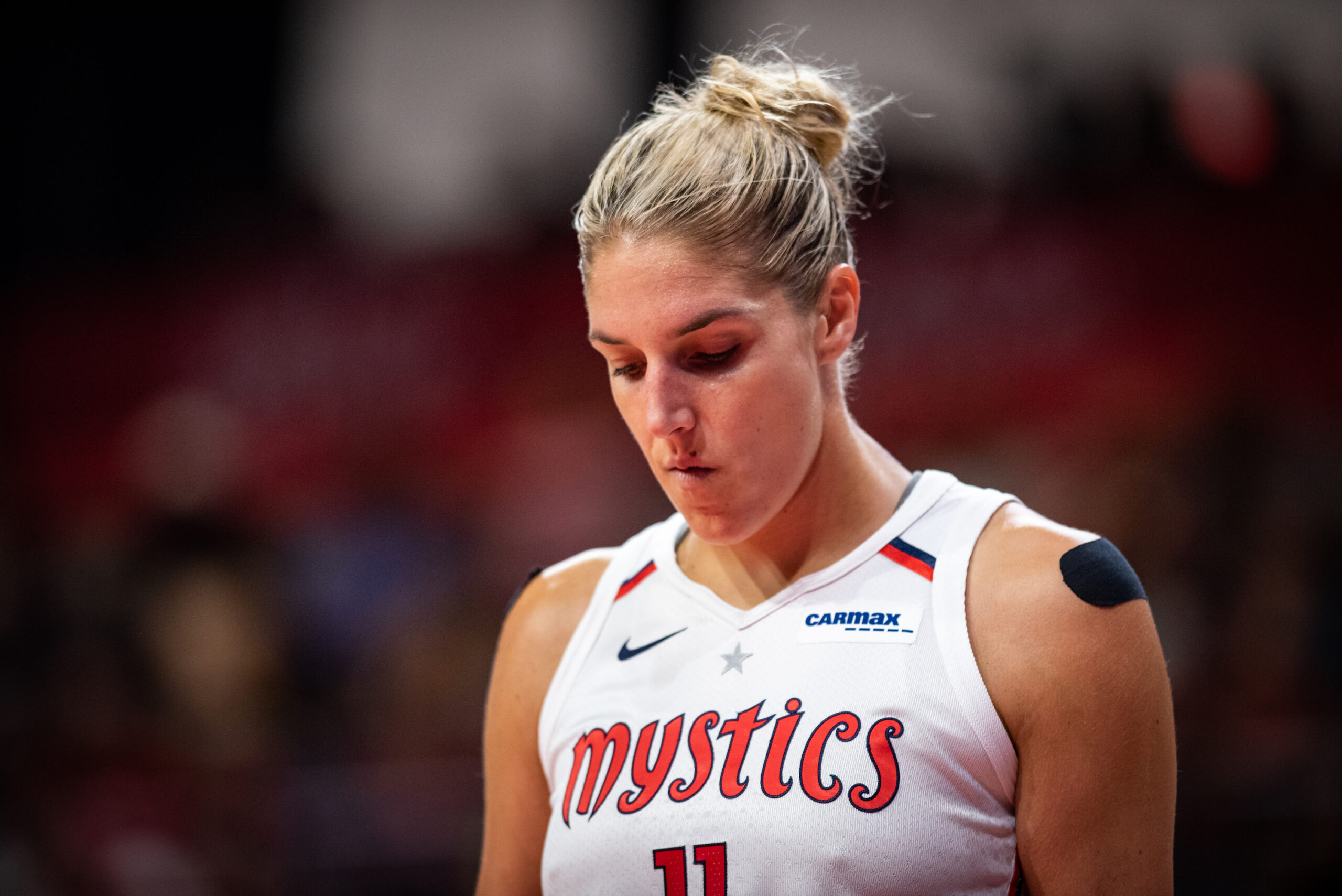 Washington Mystics forward Elena Delle Donne is shown from the chest up, wearing a white Mystics uniform and looking straight down at the ground.