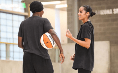 Skylar Diggins-Smith stands in a gym, talking to Bridget Pettis who has her back turned to the camera, basketball under her arm.