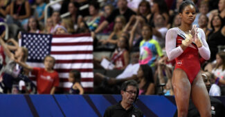 Gymnast Gabby Douglas stands with her hands clasped together as she waits to compete in the floor exercise. Behind her, a large American flag is visible in the crowd.