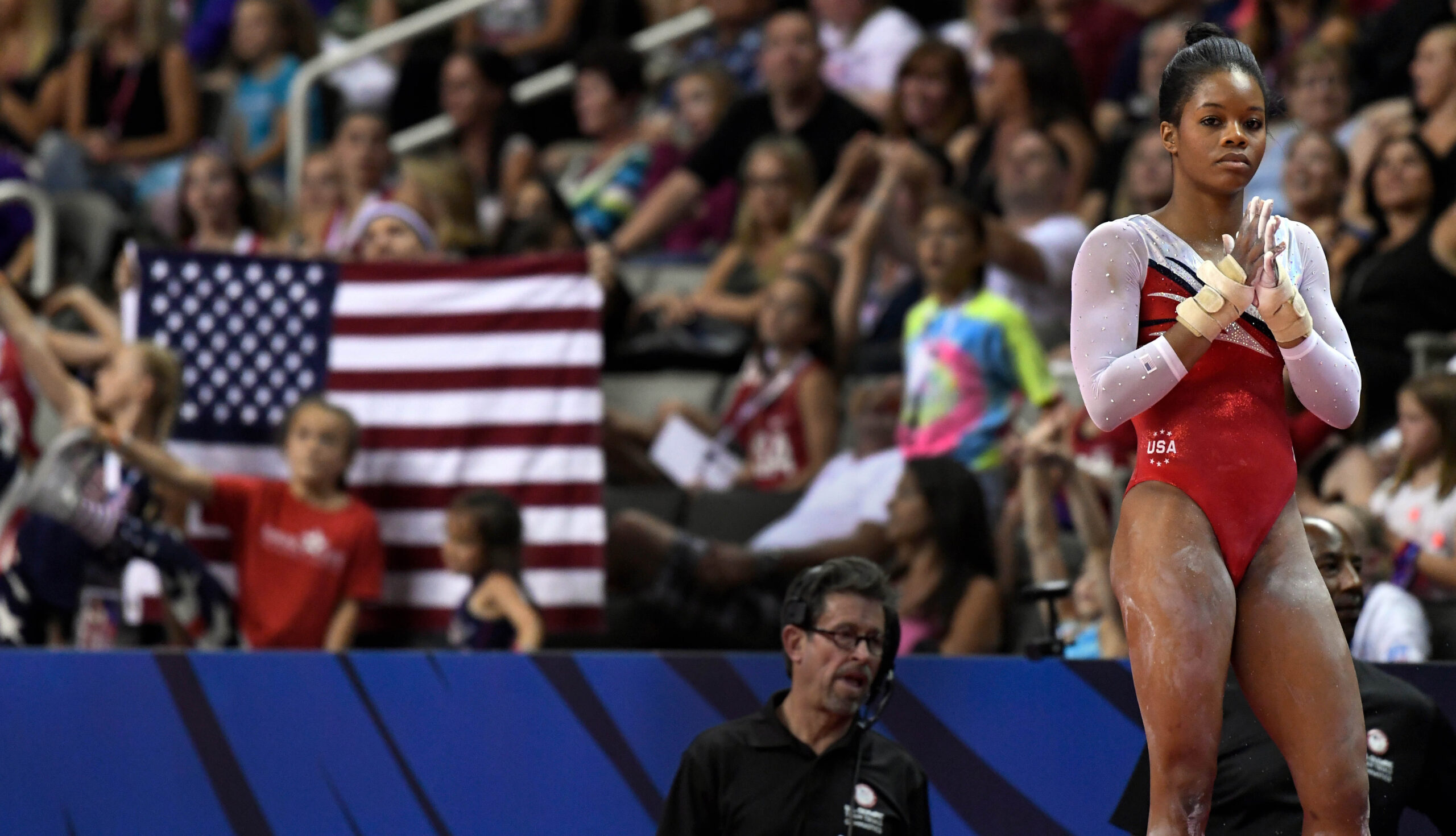 Gymnast Gabby Douglas stands with her hands clasped together as she waits to compete in the floor exercise. Behind her, a large American flag is visible in the crowd.