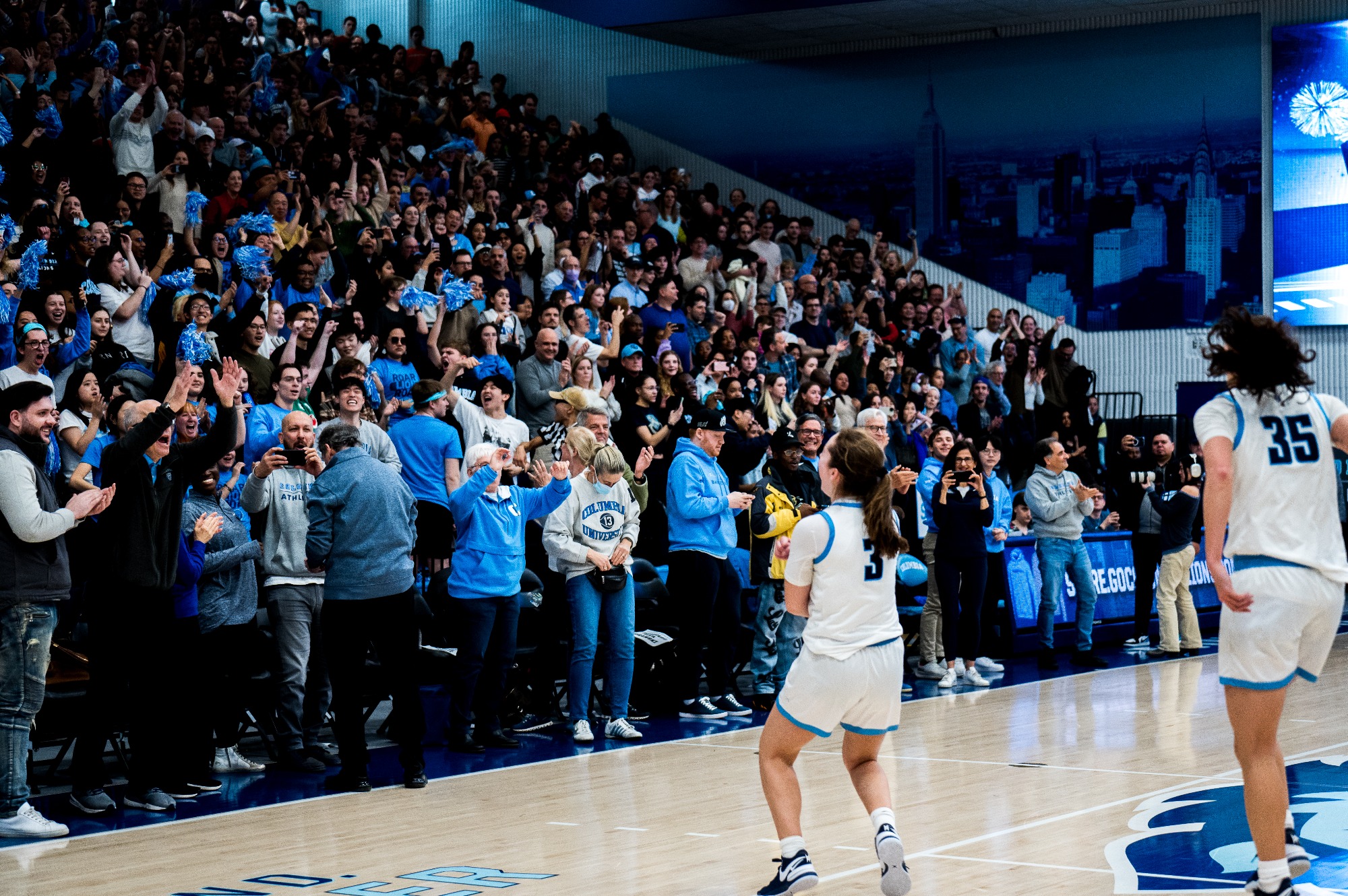 Columbia guards Cecelia Collins and Abbey Hsu celebrate on the court, facing their cheering fans in the stands.