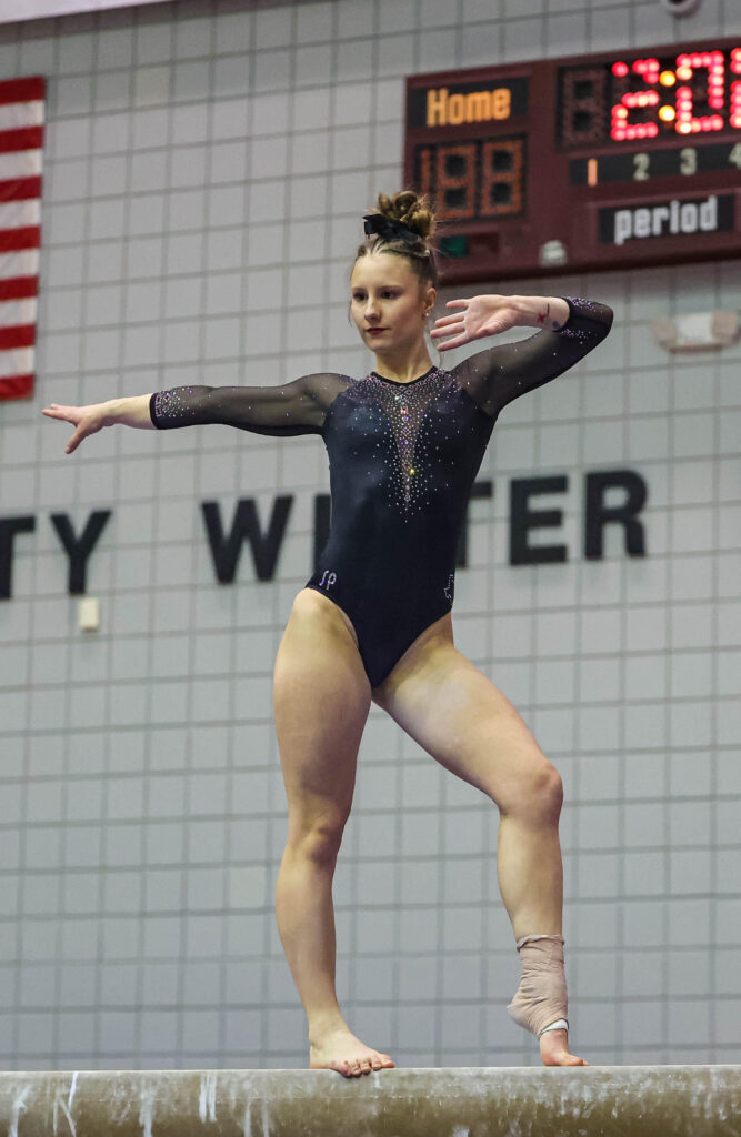 Daisy Woodring stands with two feet on the beam. One arm is fully extended to the side, and the other is bent at the elbow in front of her.