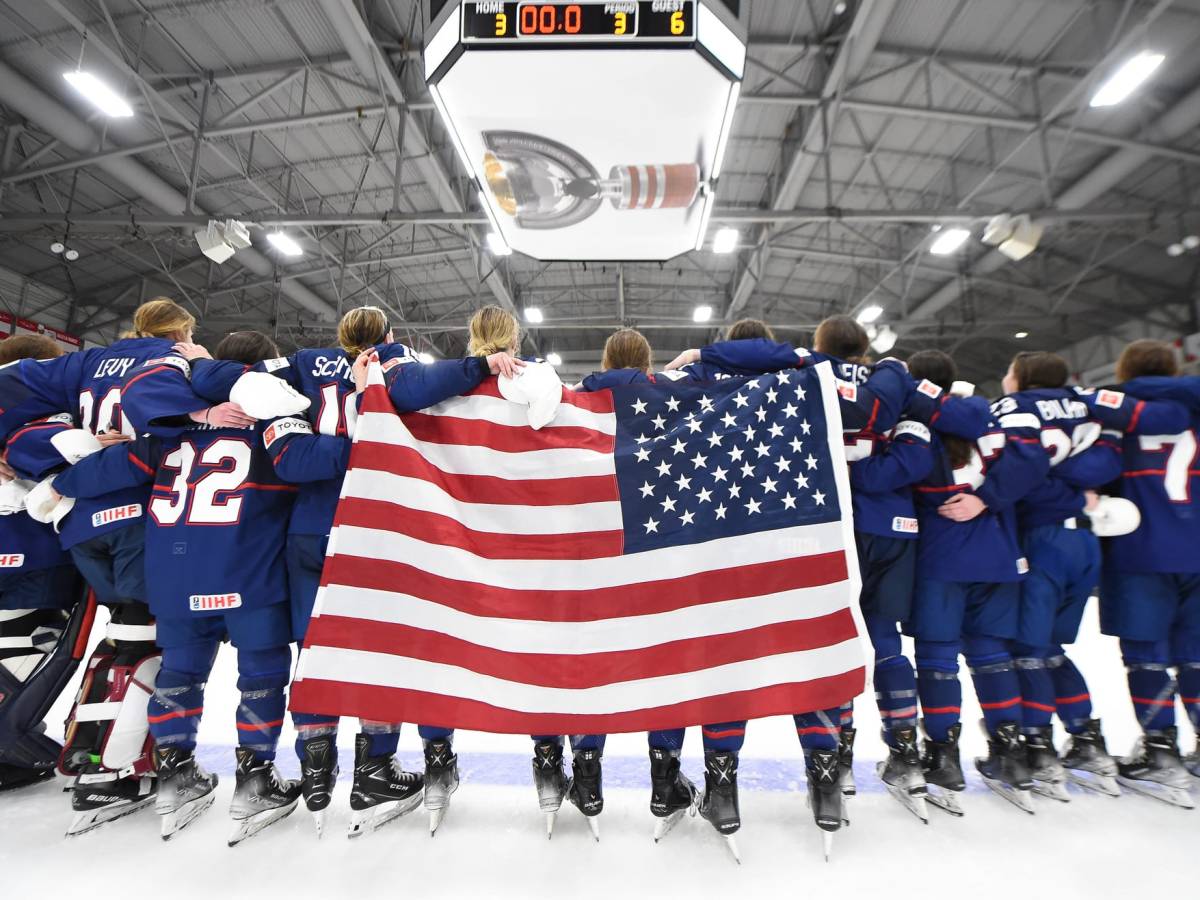 Team USA stands arm-in-arm, on the blue line. The American flag hangs over some backs.