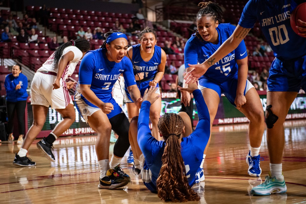Several players gather to help their teammate who is in a seated position off the ground. Another player screams while clenching their fists. In the background the head coach is clapping.