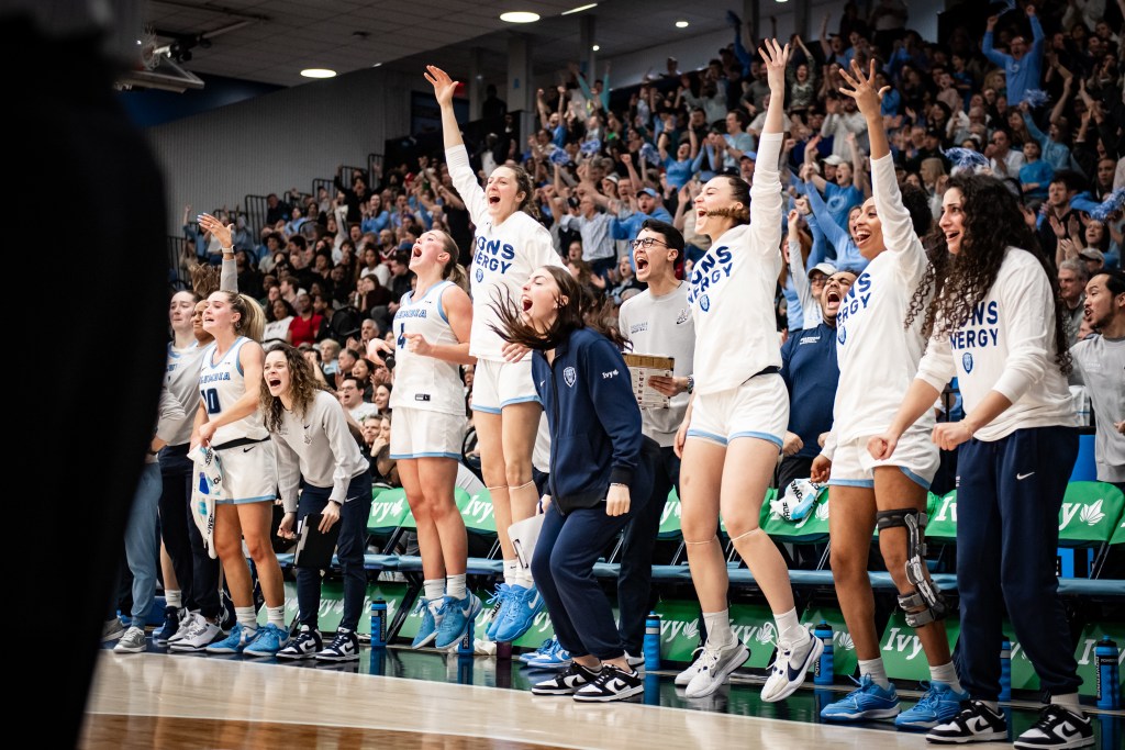Players on the Columbia bench jump up and down, shout, and raise one arm to celebrate a basket.