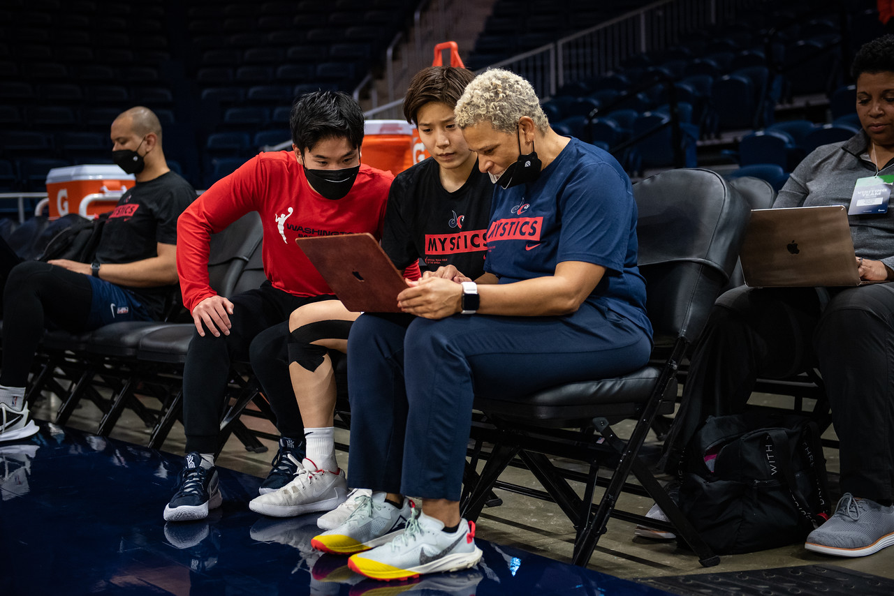 Mystics guard Rui Machida sits on the bench during practice with two assistants. They are all looking at a computer screen.