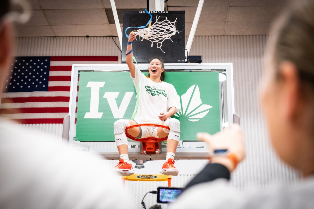 Princeton point guard Kaitlyn Chen sits on the rim after winning the Ivy League Tournament. She joyfully waves the net over her head with her right hand.