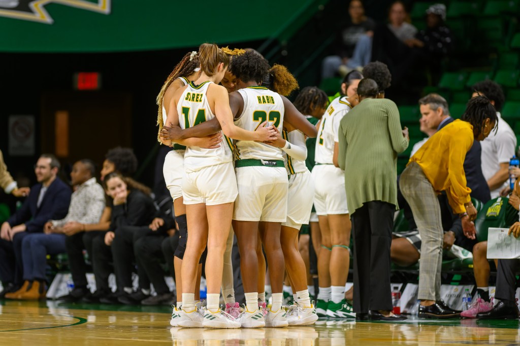 Five Mason players huddle up tightly with their arms around each other near the team's bench.