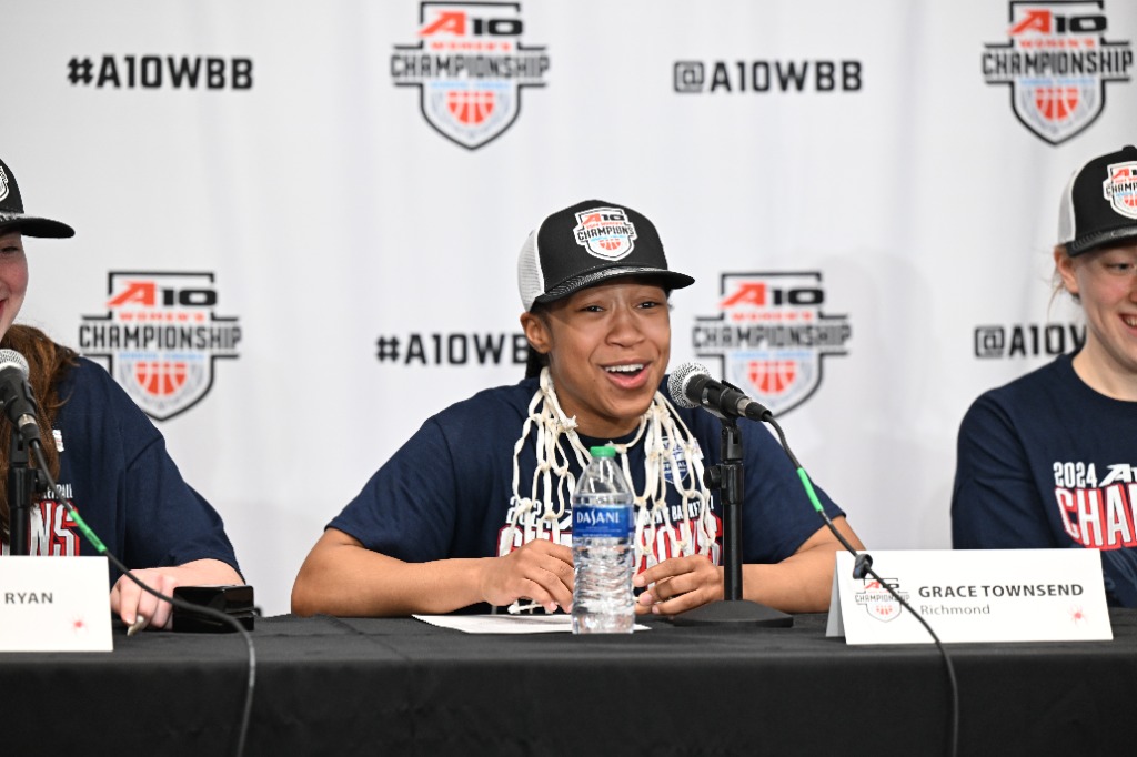 Senior guard Grace Townsend smiles with her A-10 championship hat and t-shirt on with the championship net around her neck like a necklace.