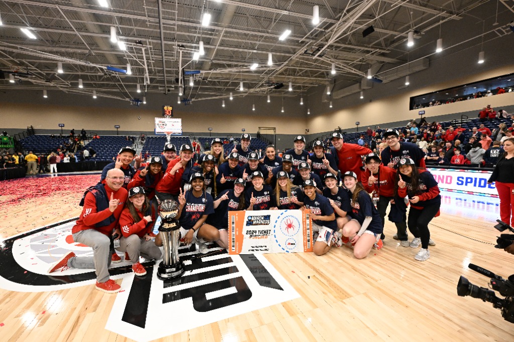 The Richmond Spiders pose with their championship hats and t-shirts with the A-10 championship trophy and newly punched giant ticket to the NCAA Tournament. Many members of the team and staff are holding up their pointer finger signaling "1."