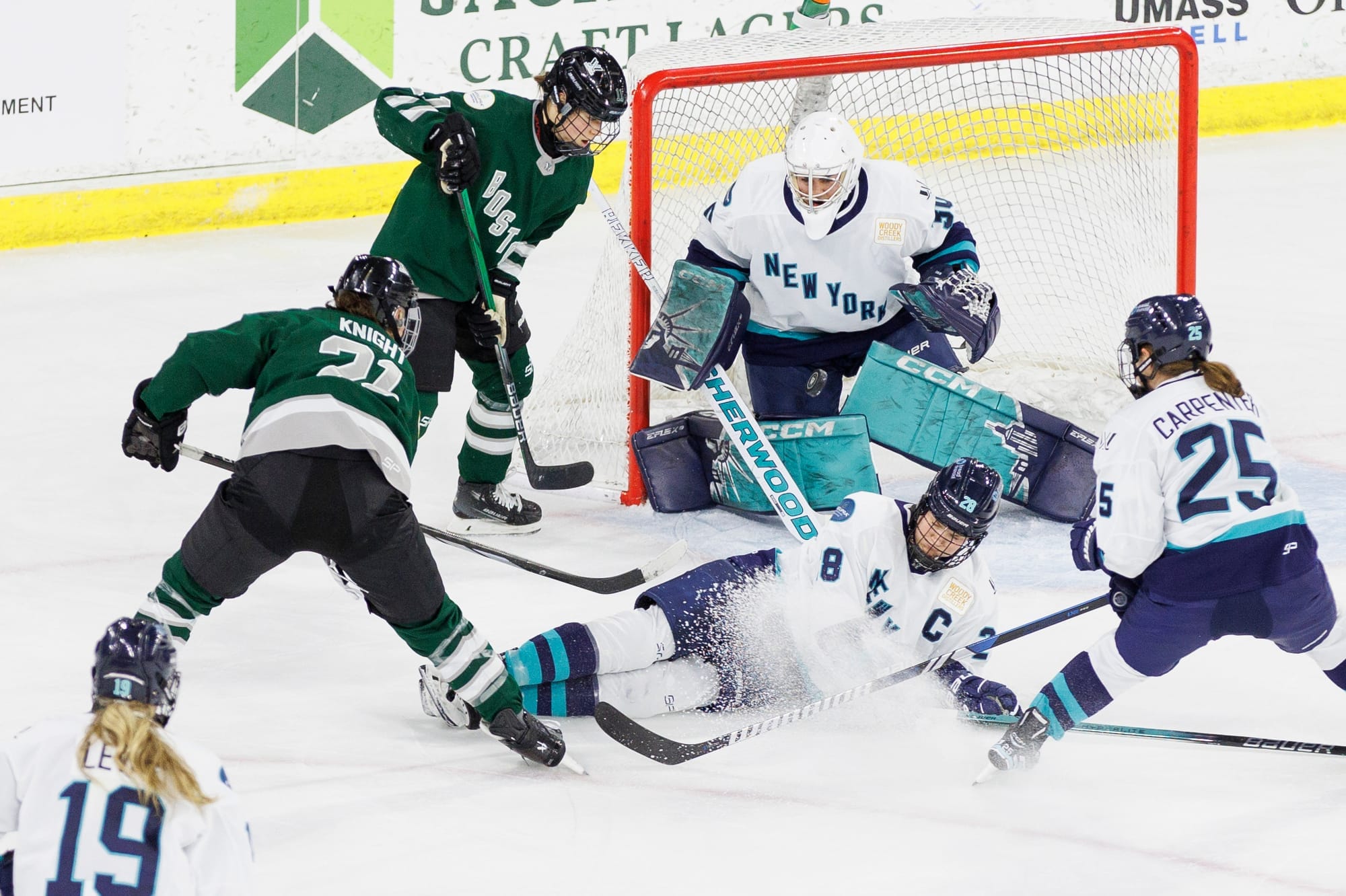 Hilary Knight takes a shot on Corinne Schroeder amidst chaos. Alina. Müller is standing off to the side of the goal. Meanwhile, New York captain Micah Zandee-Hart is laying on the ice trying to block the shot as Alex Carpenter comes to a stop. Boston players are wearing green, while New York players are in white.