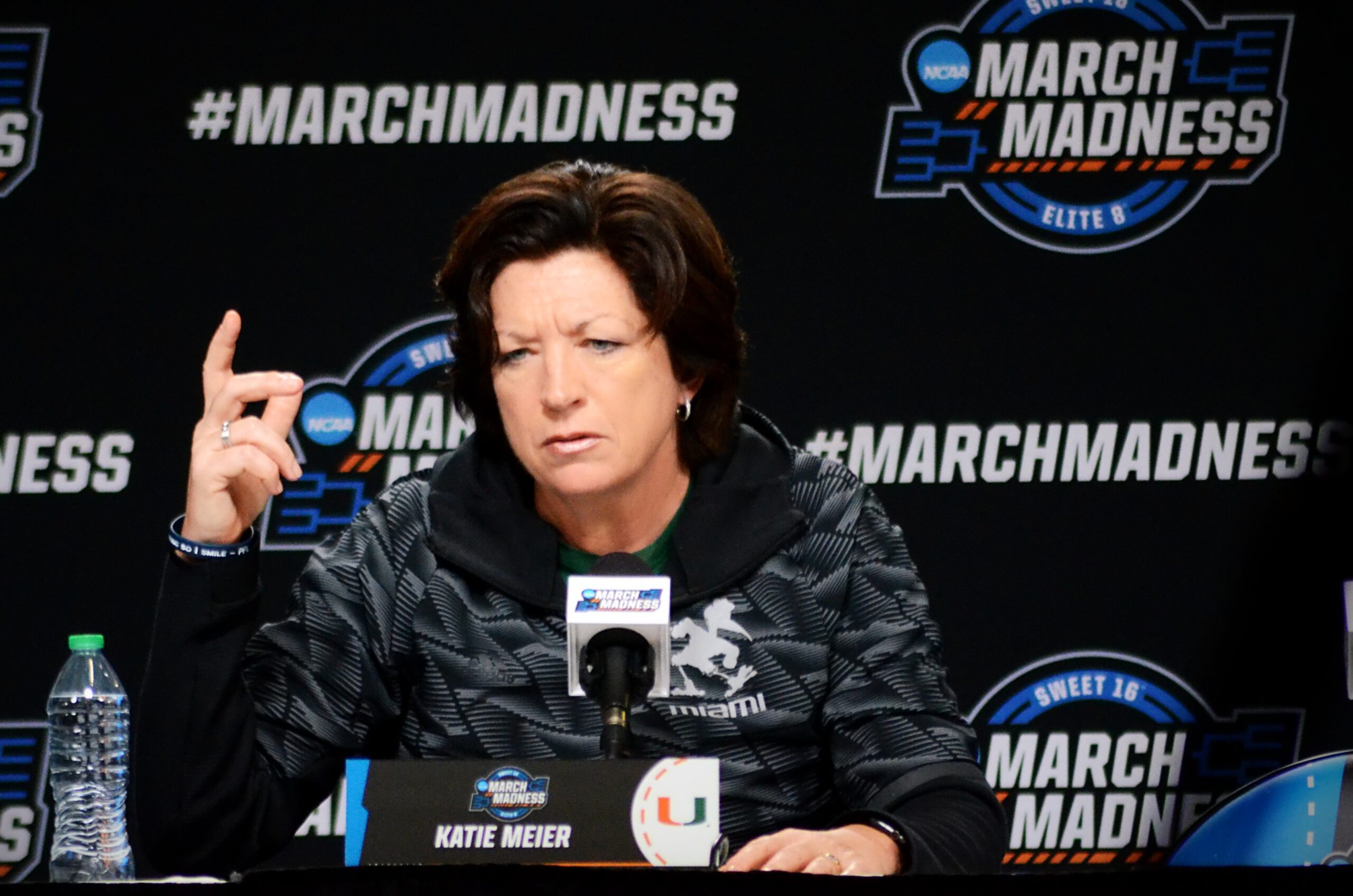 Miami head coach Katie Meier holds one finger up as she sits at the podium and speaks with reporters at the Sweet 16 in 2023. A March Madness backdrop is visible behind her.
