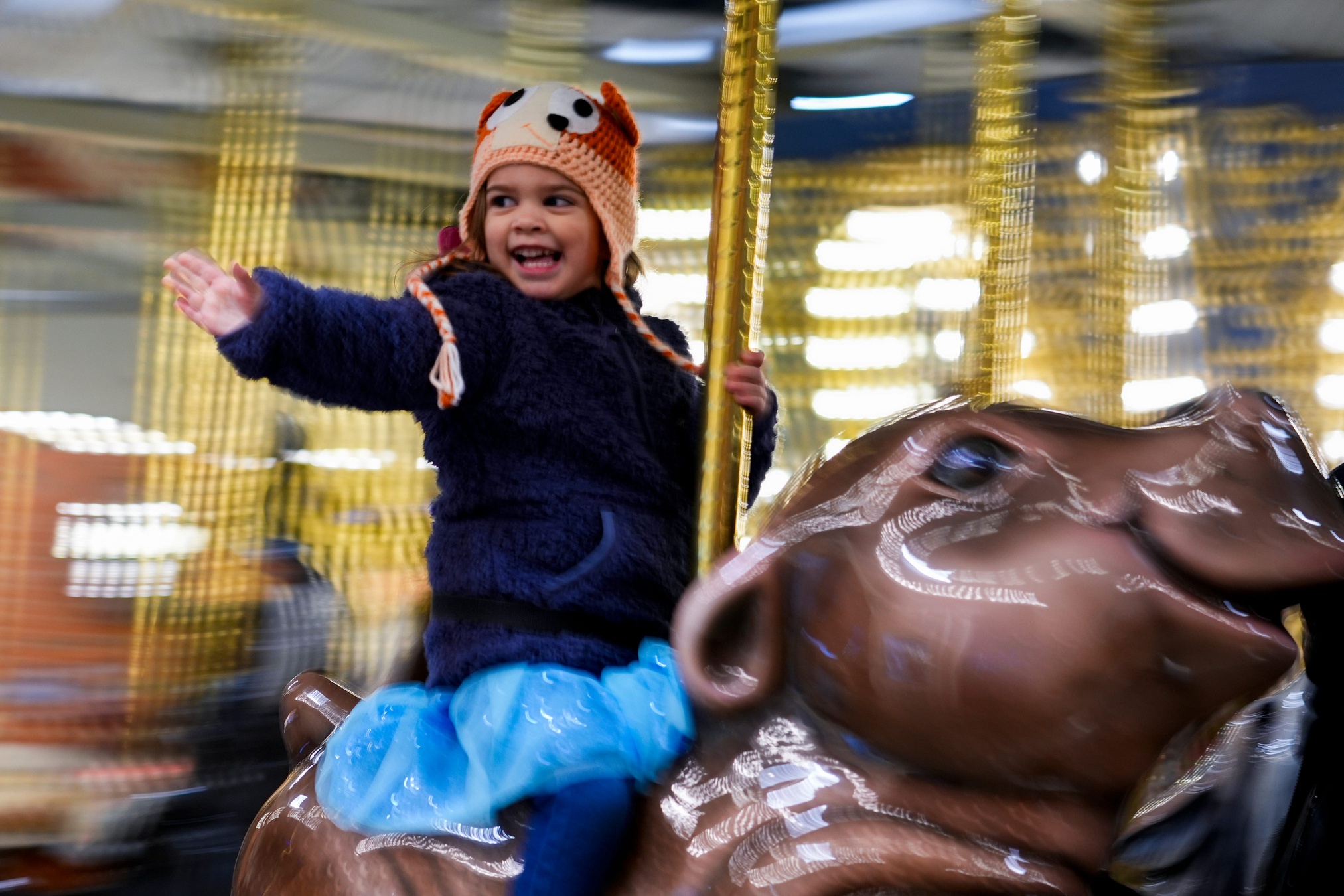 Lavender Thornhill holds on with her left hand and waves with her right as she rides a baby hippo on a carousel.