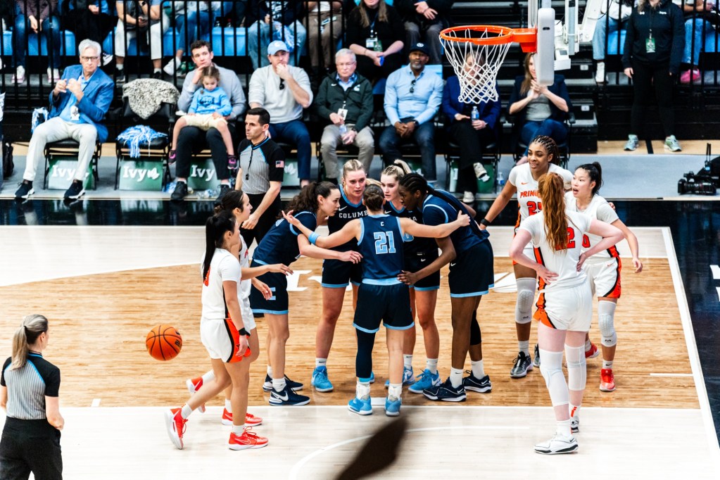 Five Columbia players come together in a huddle near the restricted arc. The five Princeton players are farther away from one another but about to gather.