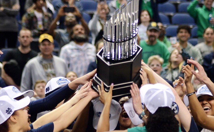 Notre Dame players and coaches form a circle with their hands around the ACC Tournament trophy.