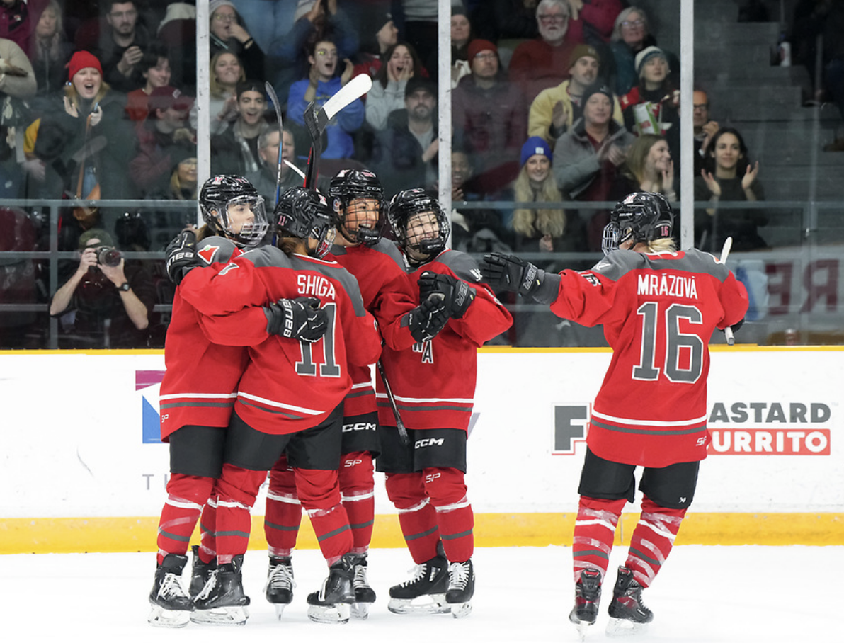Mrázová goes in to celebrate a goal with Shiga and three other Ottawa teammates. Mrázová has her arms extended for a hug as she skates towards them, while Shiga and three other players are already in a group hug. All are wearing red home uniforms.