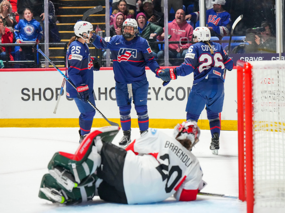 Winn is about to be in a group hug with two teammates, all wearing blue. Brändli (in white) lays on the ice in front of them.