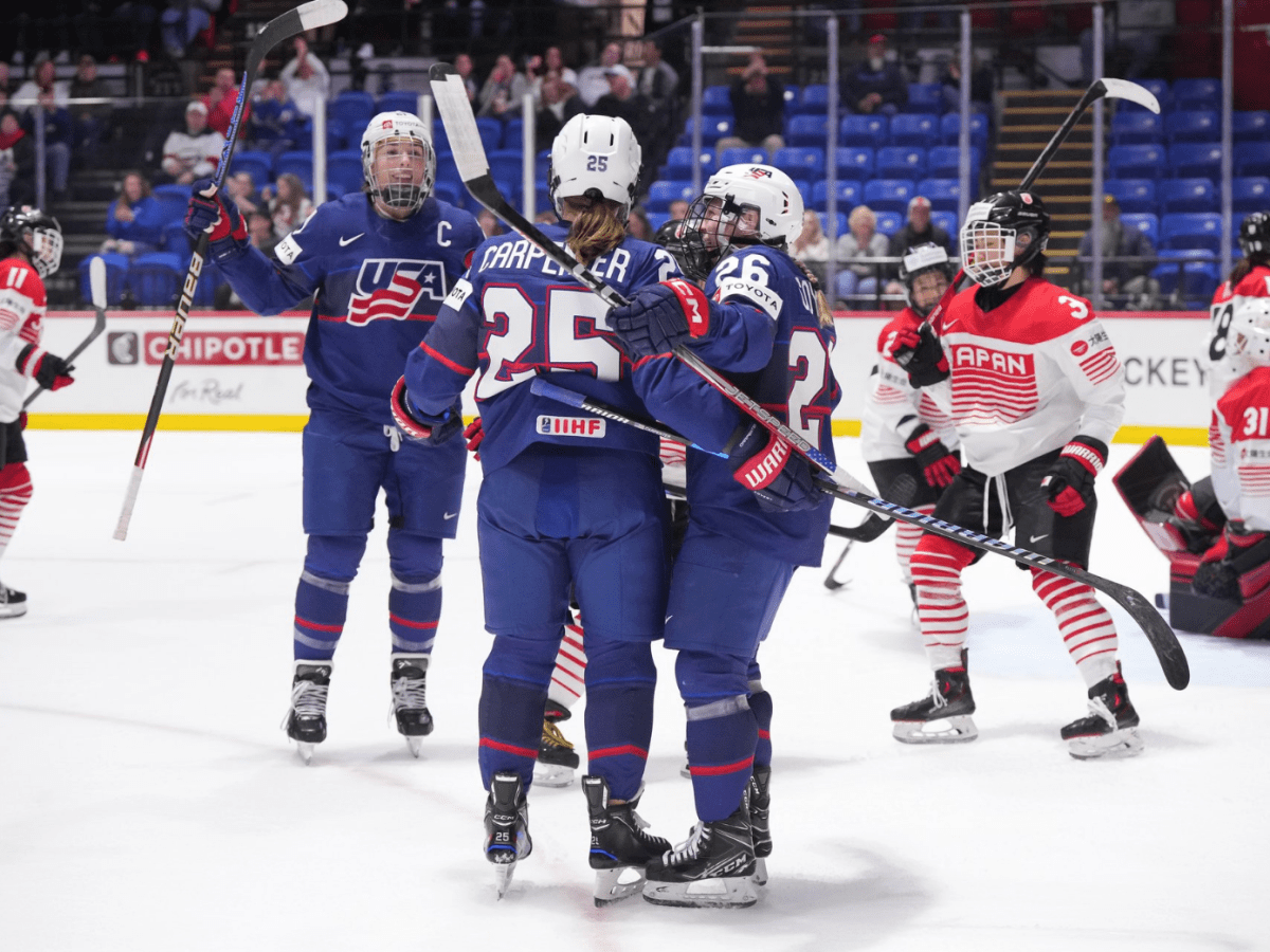 Carpenter celebrates her goal with a group hug featuring Coyne Schofield and Knight. They are wearing blue uniforms.