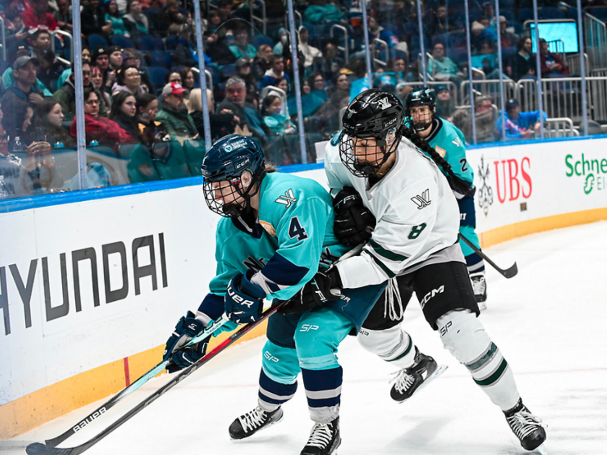 Adzija (right in white) battles for the puck with Baker (left in teal) in the corner. Adzija is reaching around Baker.