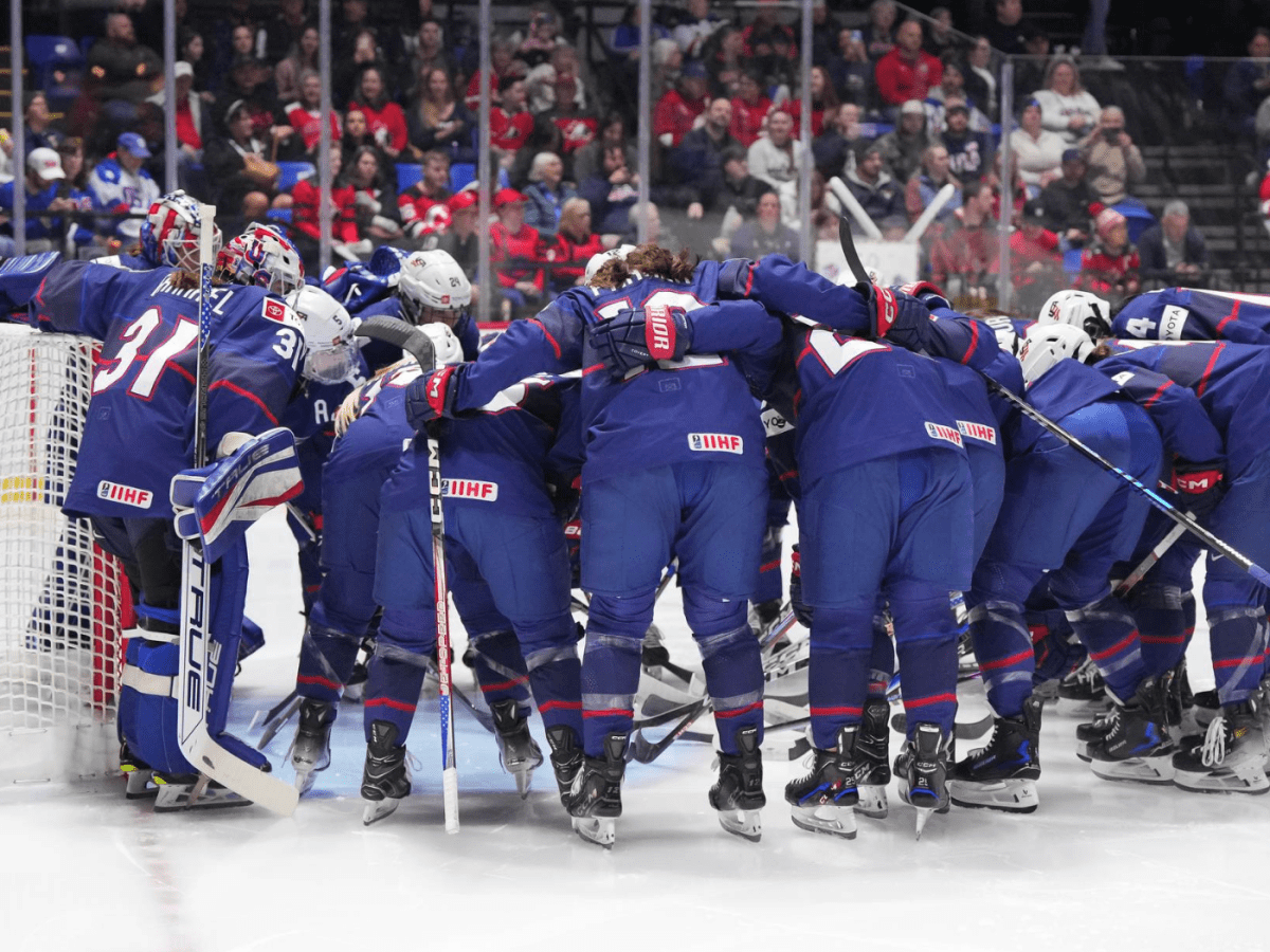 Team USA hunches over, arm-in-arm, in a huddle in front of their goal before the gold medal game. They are wearing blue.
