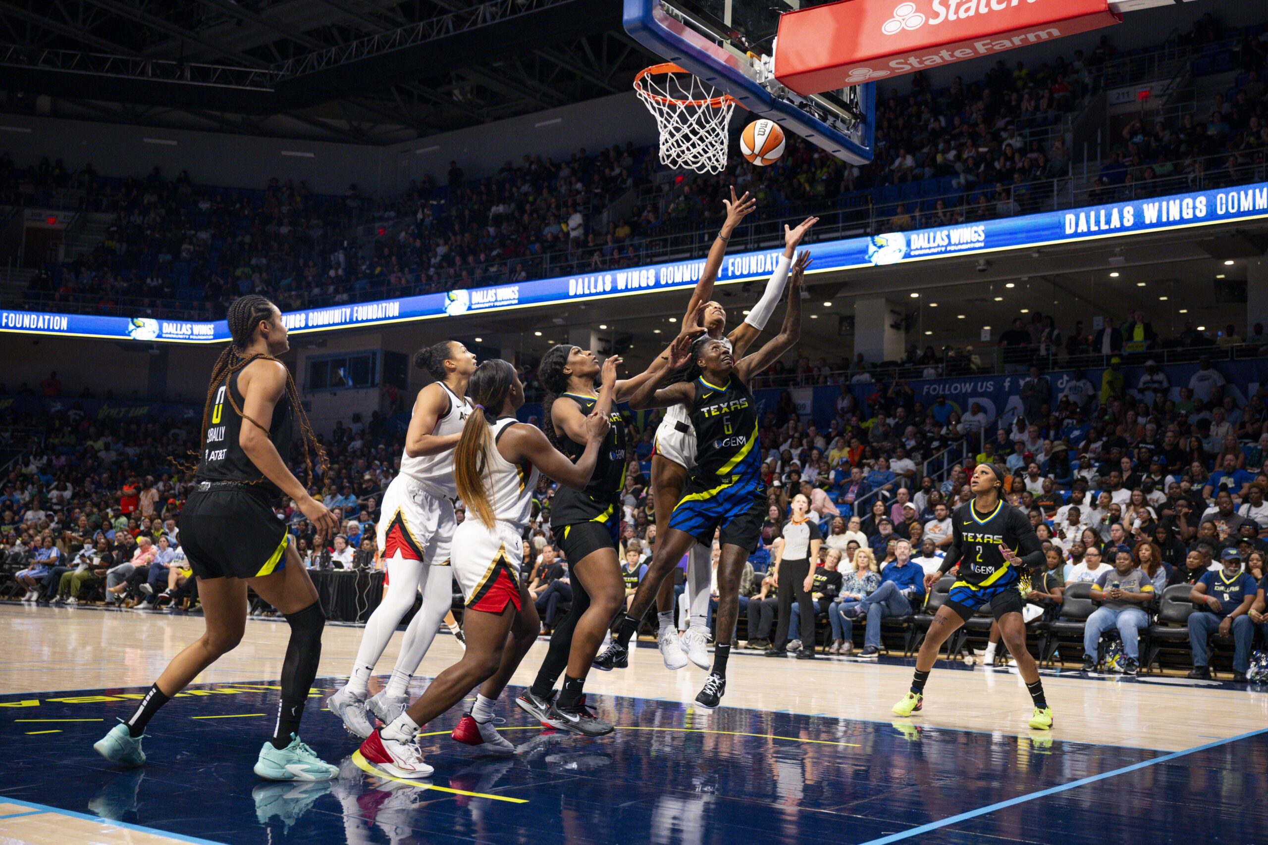 Dallas Wings and Las Vegas Aces players cluster in the lane in pursuit of a rebound.