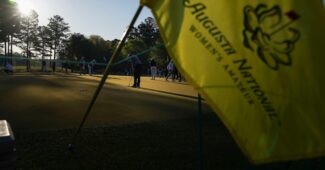 A yellow flag for the Augusta National Women's Amateur is shown in the foreground. Golfers warm up on the practice green in the background. The sun is low in the sky, casting shadows on much of the green.