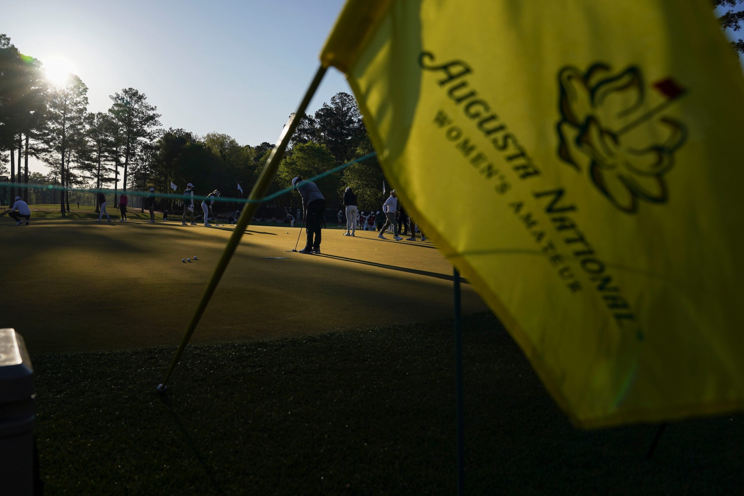 A yellow flag for the Augusta National Women's Amateur is shown in the foreground. Golfers warm up on the practice green in the background. The sun is low in the sky, casting shadows on much of the green.