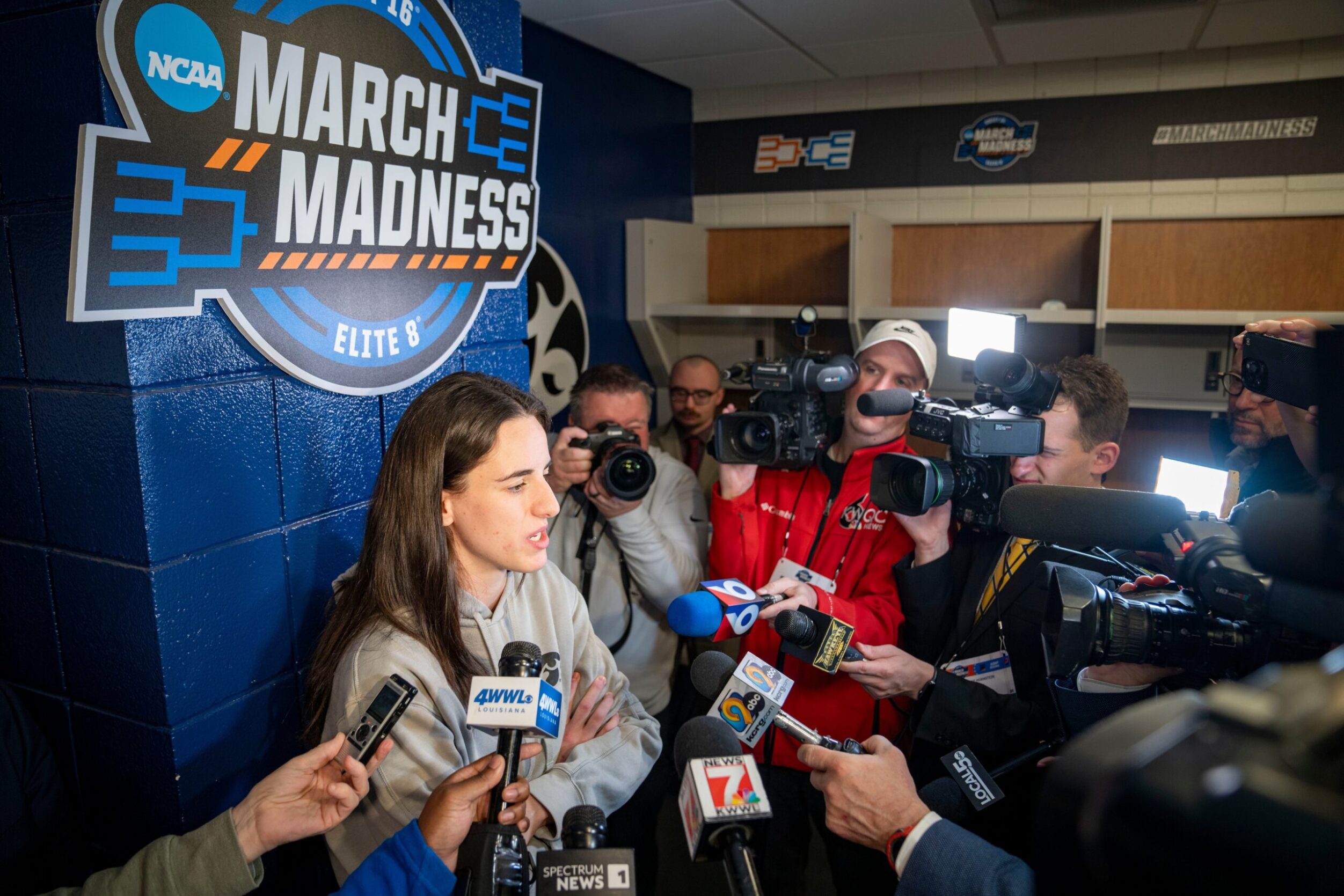 Caitlin Clark stands against a wall surrounded by media members with outstretched microphones