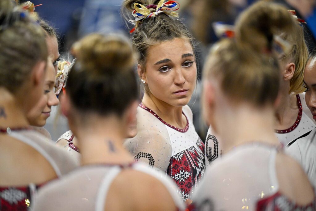A close-up photo of Oklahoma gymnasts huddling up. Most of their faces aren't visible, but one gymnast facing the camera looks somber.