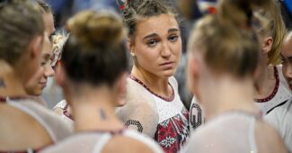 A close-up photo of Oklahoma gymnasts huddling up. Most of their faces aren't visible, but one gymnast facing the camera looks somber.