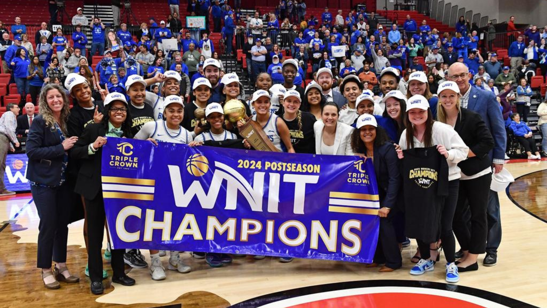 Saint Louis players, coaches and staff gather to take a photo with the purple 2024 Postseason WNIT Champions banner. Most are wearing white hats and either wearing or holding up black championship shirts. One player is also holding the trophy with a smile.