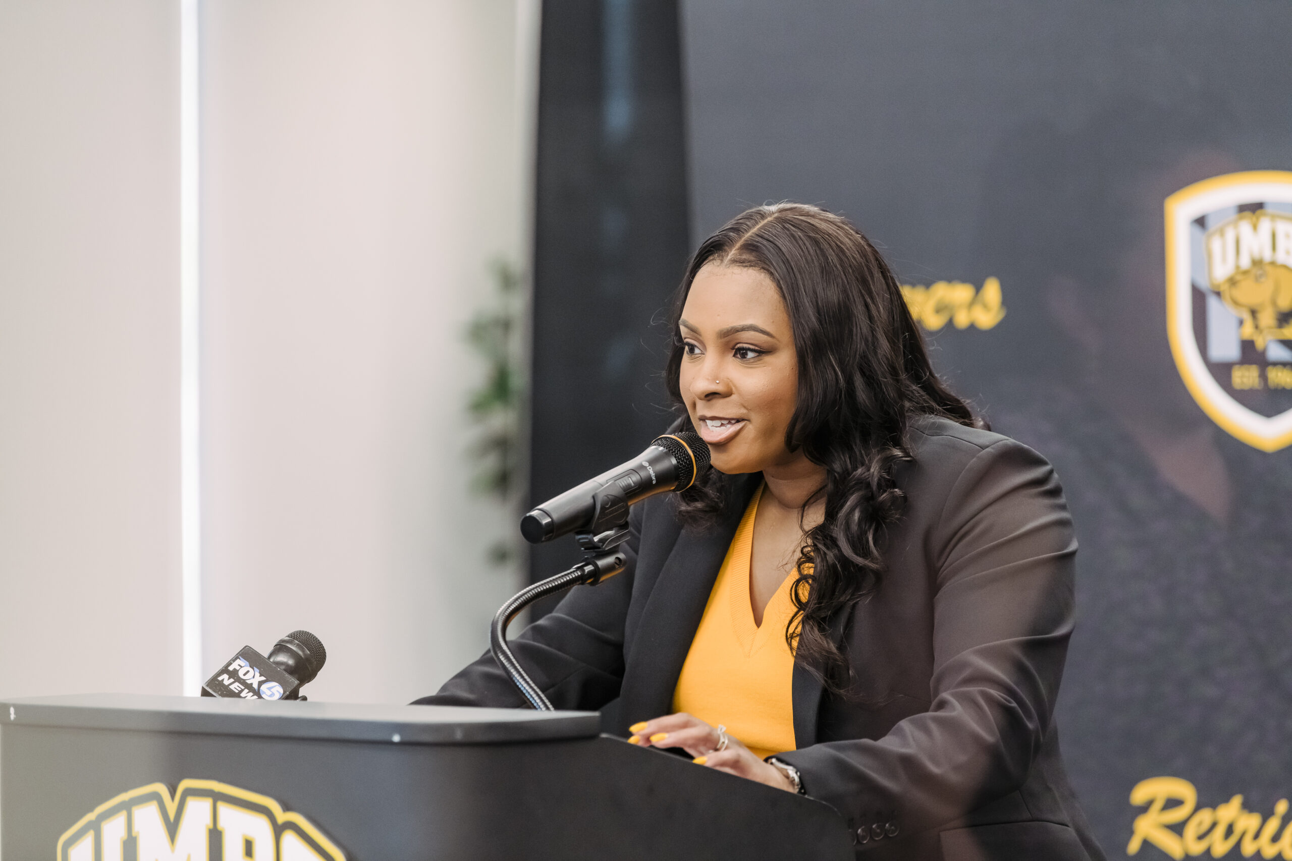 UMBC head coach Candice Hill leans into the microphone with both hands during her introductory press conference. She's wearing a "Retriever gold" shirt and black blazer, matching the school's colors.