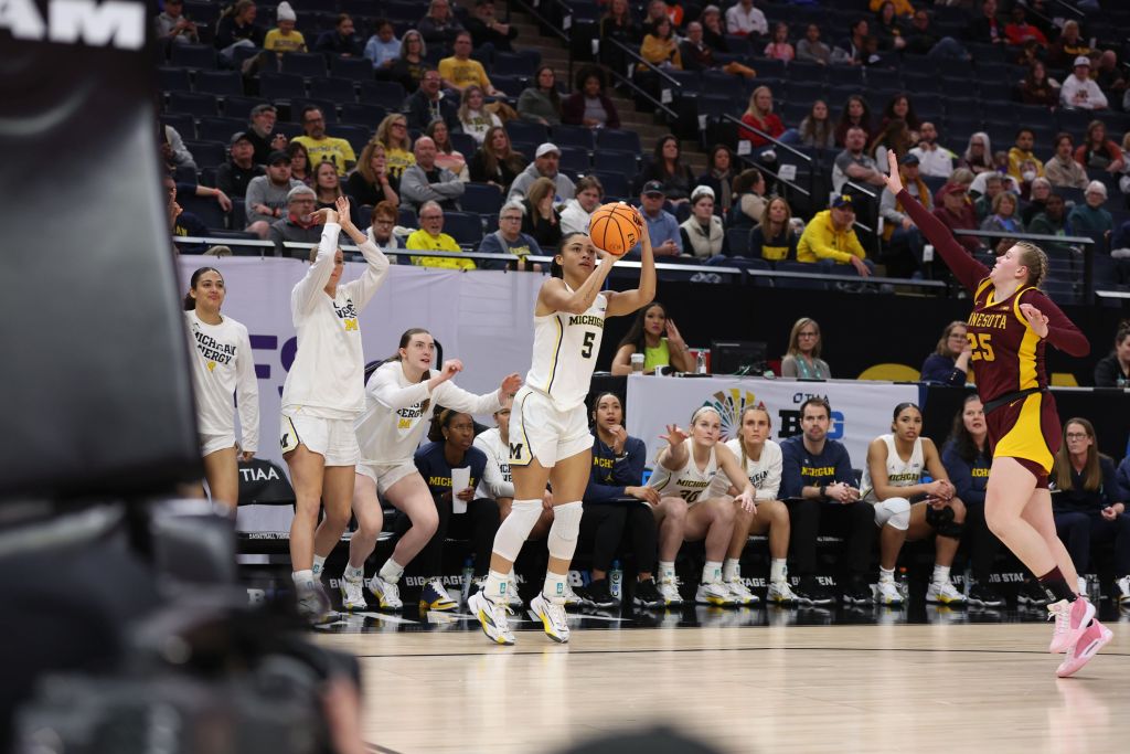 Several players on the Michigan bench rise as teammate Laila Phelia shoots a 3-pointer right in front of them. The coaching staff, including Ariel Atkins, stays seated but watches intently.
