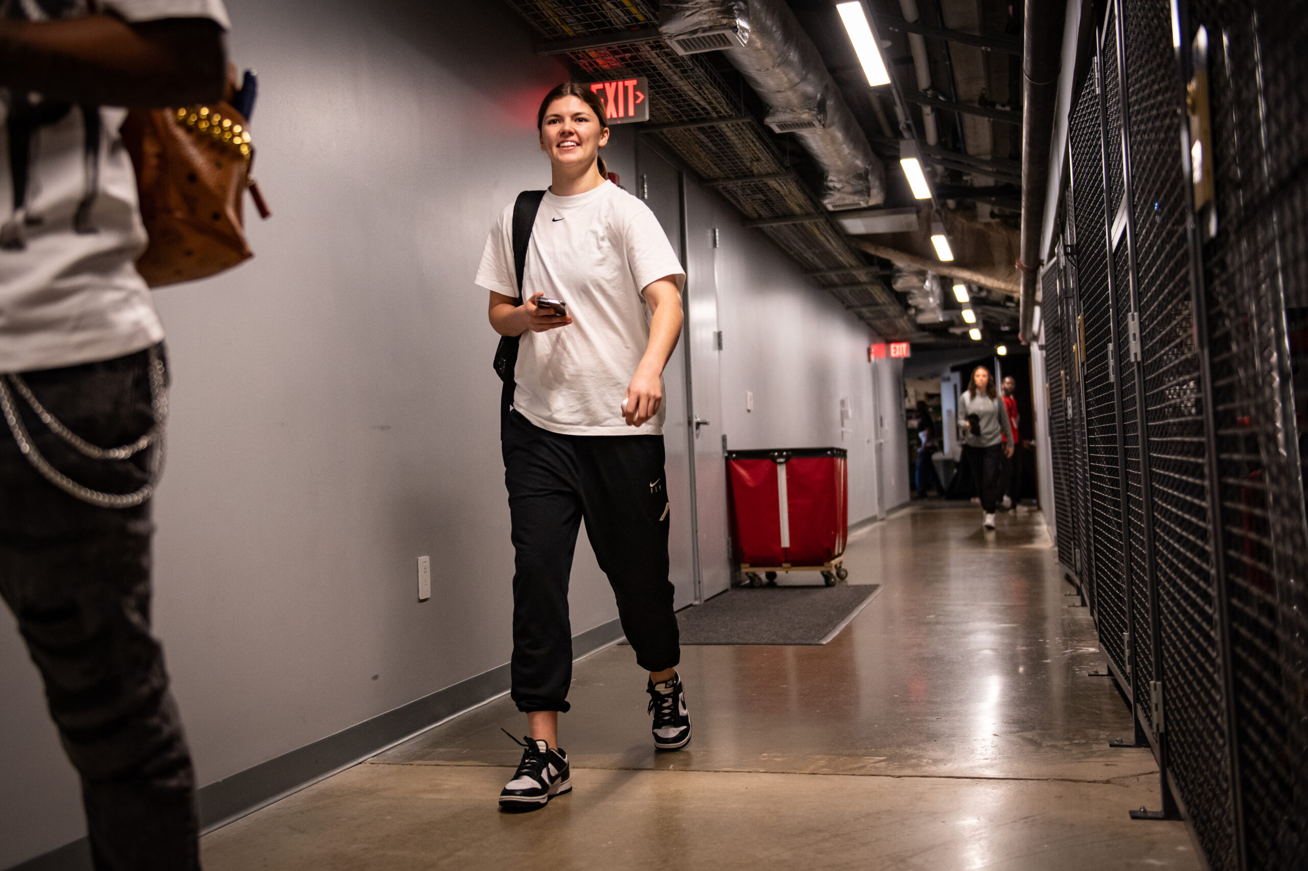 Seattle Storm guard Jade Melbourne walks down a hallway at the Entertainment and Sports Arena. She is wearing a white T-shirt and black pants and has a backpack slung over her right shoulder.