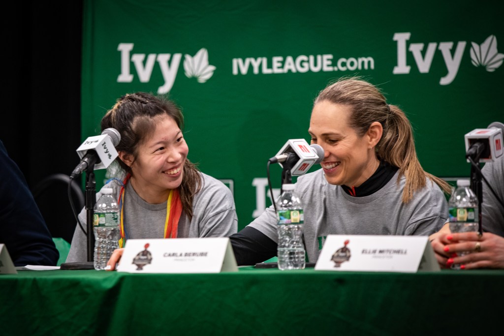 Princeton guard Kaitlyn Chen and head coach Carla Berube sit at the podium for a postgame press conference. Berube grins and looks down at the table, while Chen looks at Berube and smiles.