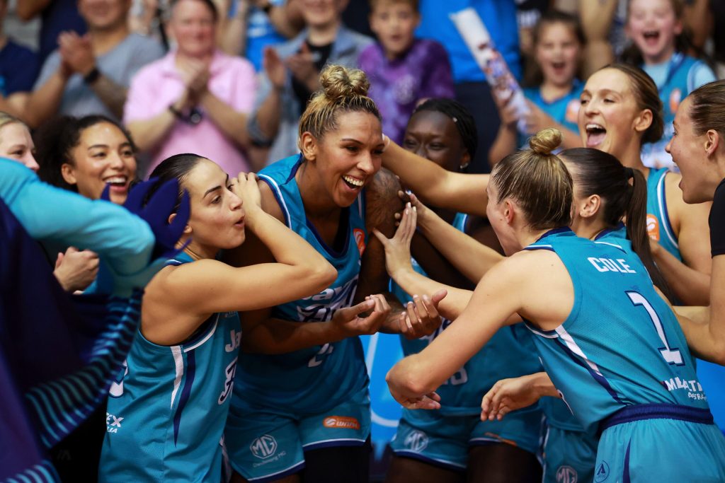 Southside Flyers players surround Russell, patting her on the back and pushing her forward to accept the WNBL Grand Final series MVP award. Russell shrinks into herself while smiling widely.