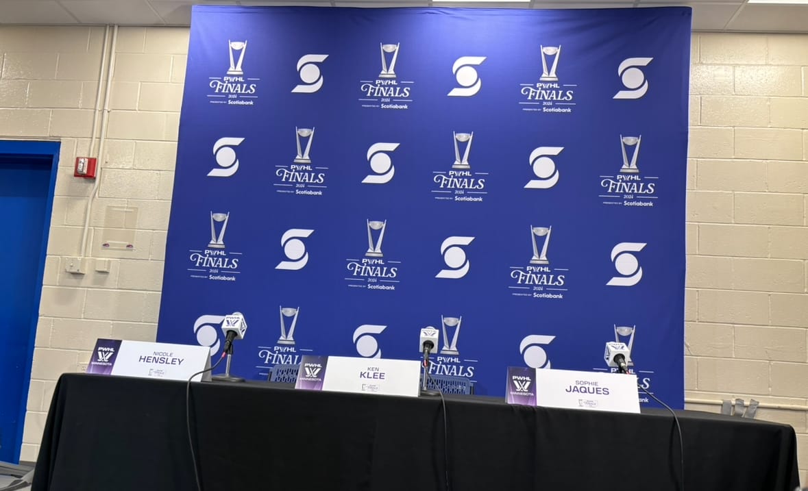 A purple backdrop with the Scotiabank and PWHL Finals logos alternating on it is sitting behind a table covered by a black tablecloth. On the table is three microphones, next to which are signs reading "Nicole Hensley," "Ken Klee," and "Sophie Jaques."