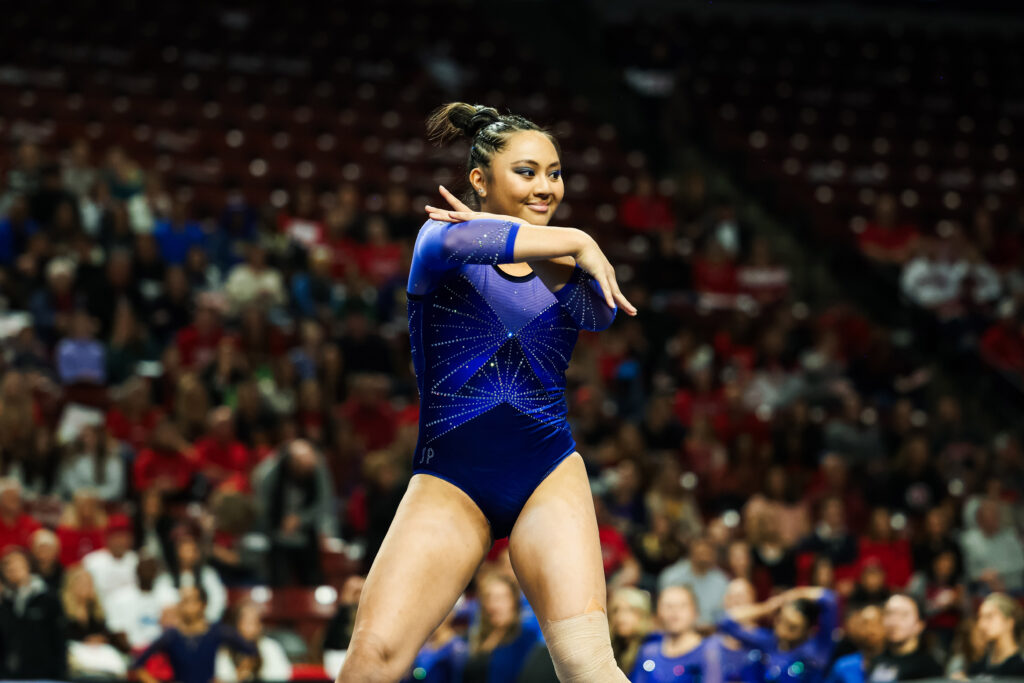 A gymnast in a blue leotard performs on the floor exercise mat.