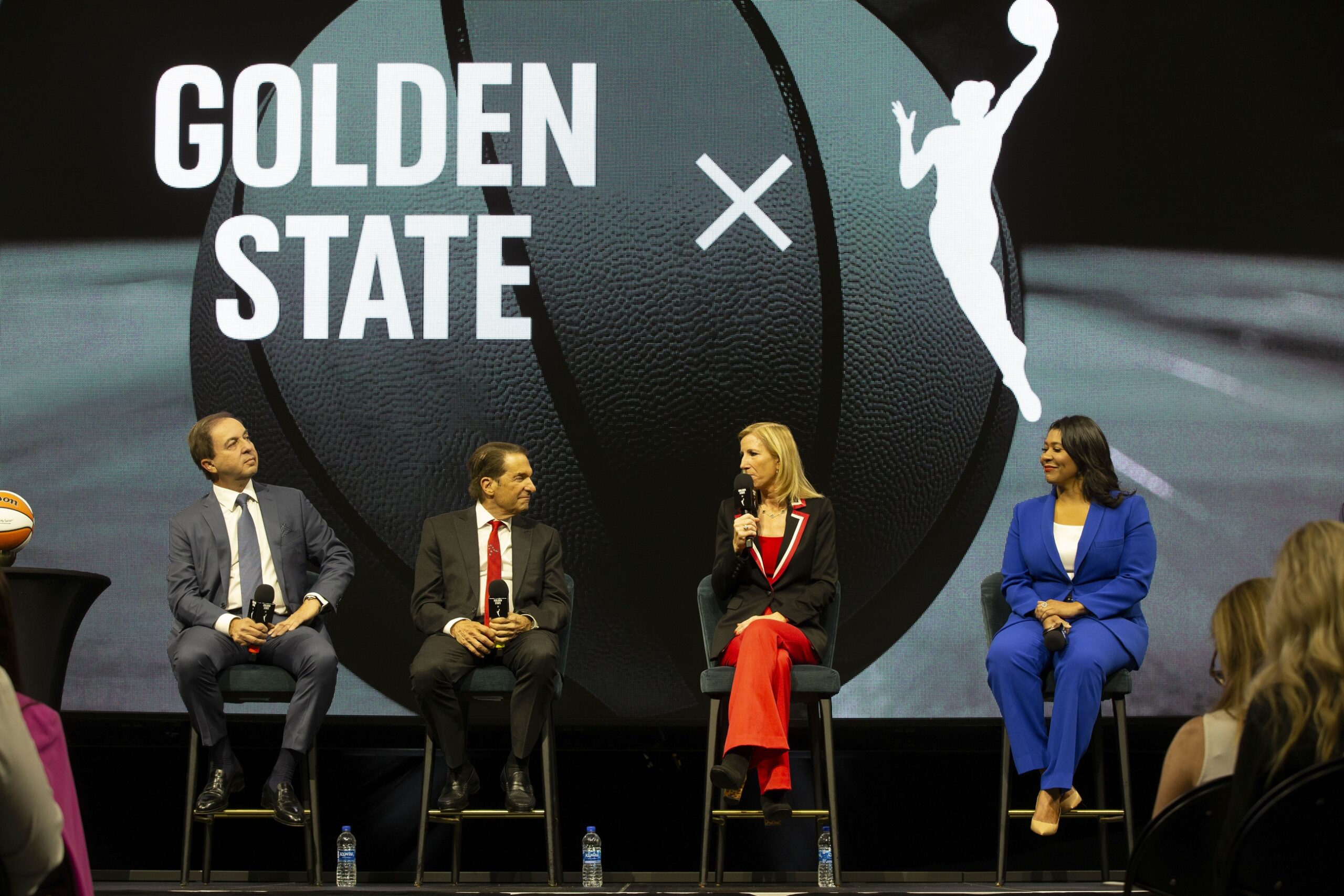 WNBA Commissioner Cathy Engelbert and a panel of guests speak in front of a large screen announcing the WNBA Golden State expansion franchise. The screen has a black-and-white photo of a basketball with the text "Golden State," an "x," and the WNBA's logo in white.