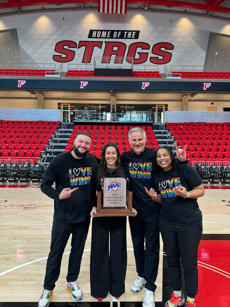 The Fairfield coaching staff poses for a photo with the MAAC regular-season championship trophy. They are all wearing black shirts that read, "Love wins" in rainbow colors.