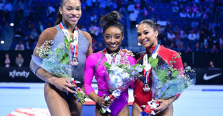 Three gymnasts in leotards, carrying flowers and wearing medals, stand on the podium at the Core Hydration Classic.