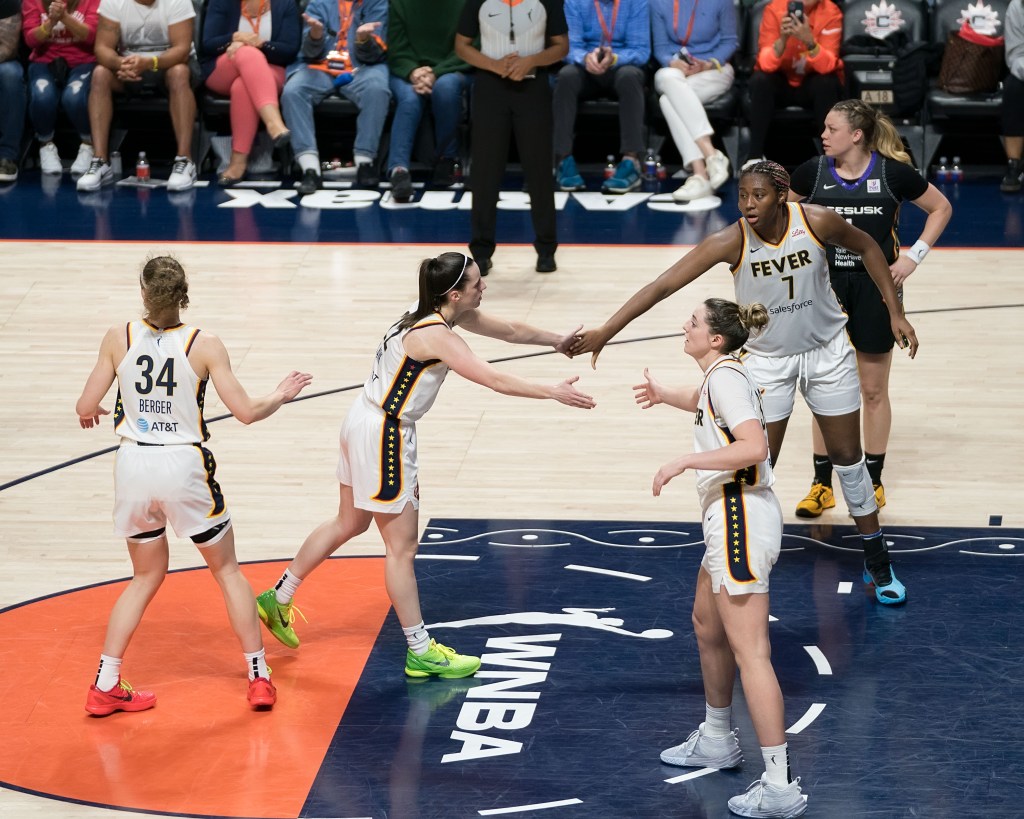 Indiana Fever wing Katie Lou Samuelson and forward/center Aliyah Boston stand in the lane and high-five guard Caitlin Clark, who is at the free-throw line. Guard Grace Berger is behind Clark waiting to congratulate her.