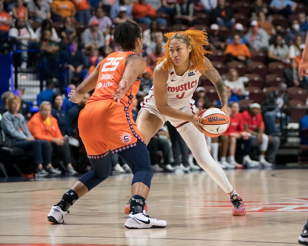 Washington Mystics center/forward Shakira Austin holds the ball with two hands near her left hip. Her feet are spread wide and she looks ahead to her right. Connecticut Sun forward Alyssa Thomas has her arms out as she defends.