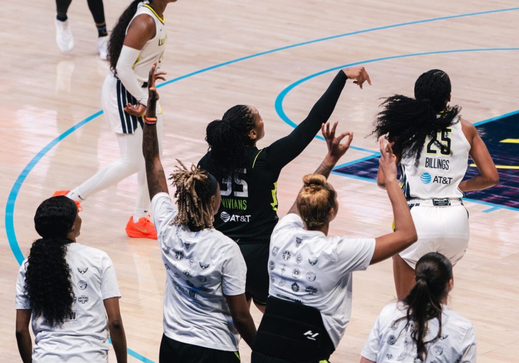 Victoria Vivians holds up her arm at the end of her three-point shooting motion and her teammates on the bench raise their arms in expectation of a make behind her.