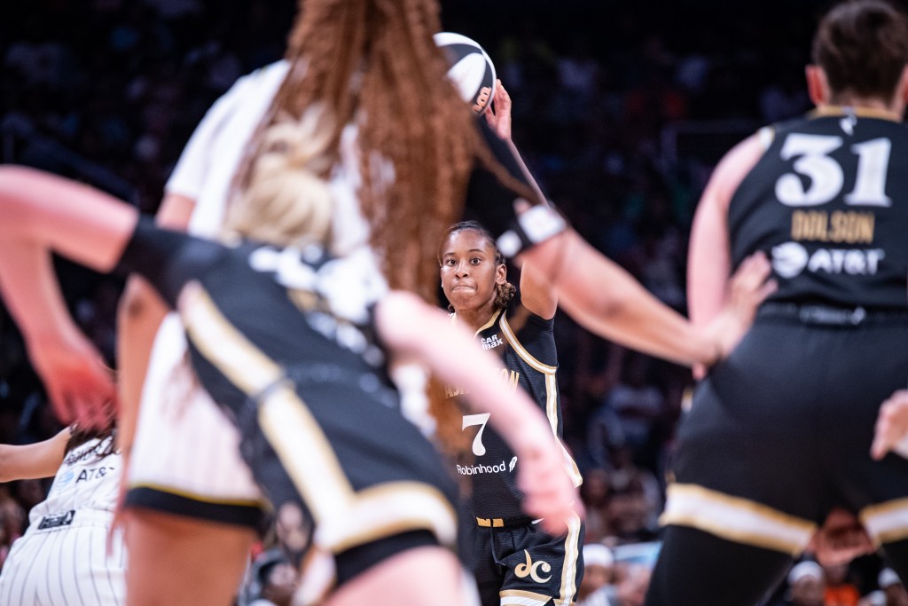 Washington Mystics guard Ariel Atkins holds the ball and looks for a pass. She is shown in the background but in focus, with several other players in the foreground with their backs to the camera.