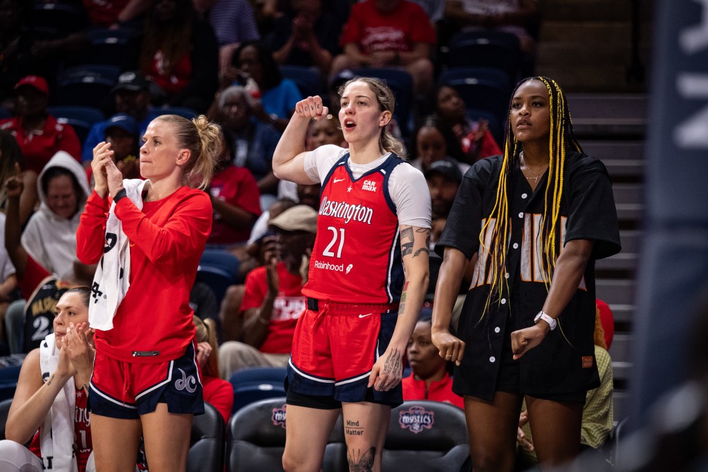 On the Washington Mystics bench, point guard Julie Vanloo claps both hands, forward Emily Engstler flexes one arm up and forward Aaliyah Edwards flexes both arms down in celebration. Vanloo and Engstler are in uniform, but Edwards is in street clothes.