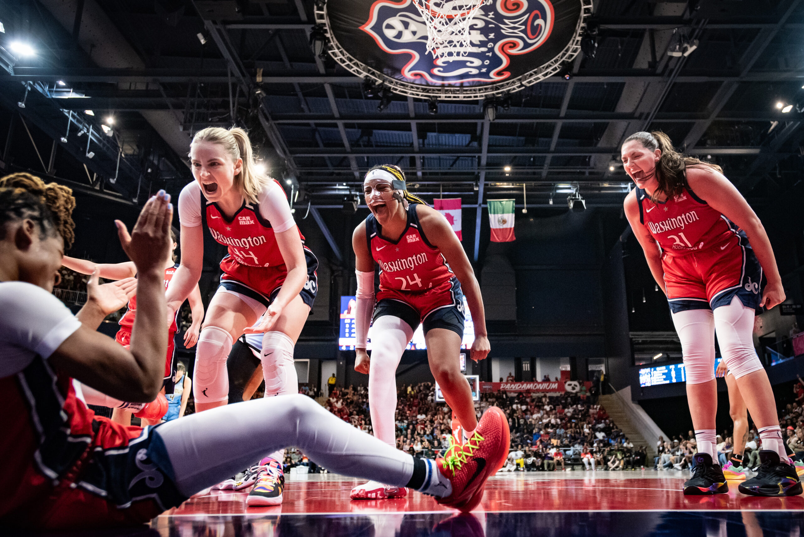 Washington Mystics guard Ariel Atkins sits on the court and claps her hands. Teammates Karlie Samuelson, Aaliyah Edwards and Stefanie Dolson yell in celebration, and Samuelson extends her hands to help Atkins up.