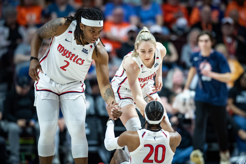 Washington Mystics forward Myisha Hines-Allen and wing Karlie Samuelson each reach one hand out to help up point guard Brittney Sykes, who is facing them with her back to the camera.