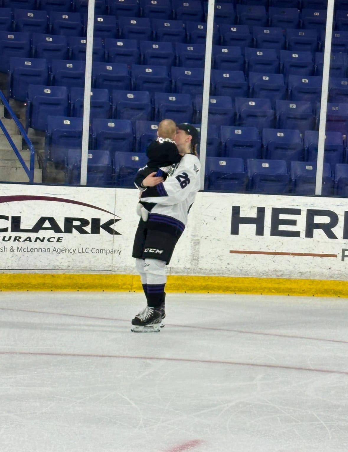 Kendall Coyne Schofield skates in the corner while holding her son, Drew, who is back-to the camera wearing a big "Walter Cup champion" chain and a black sweatsuit. She is wearing the same outfit as the press scrum photo.