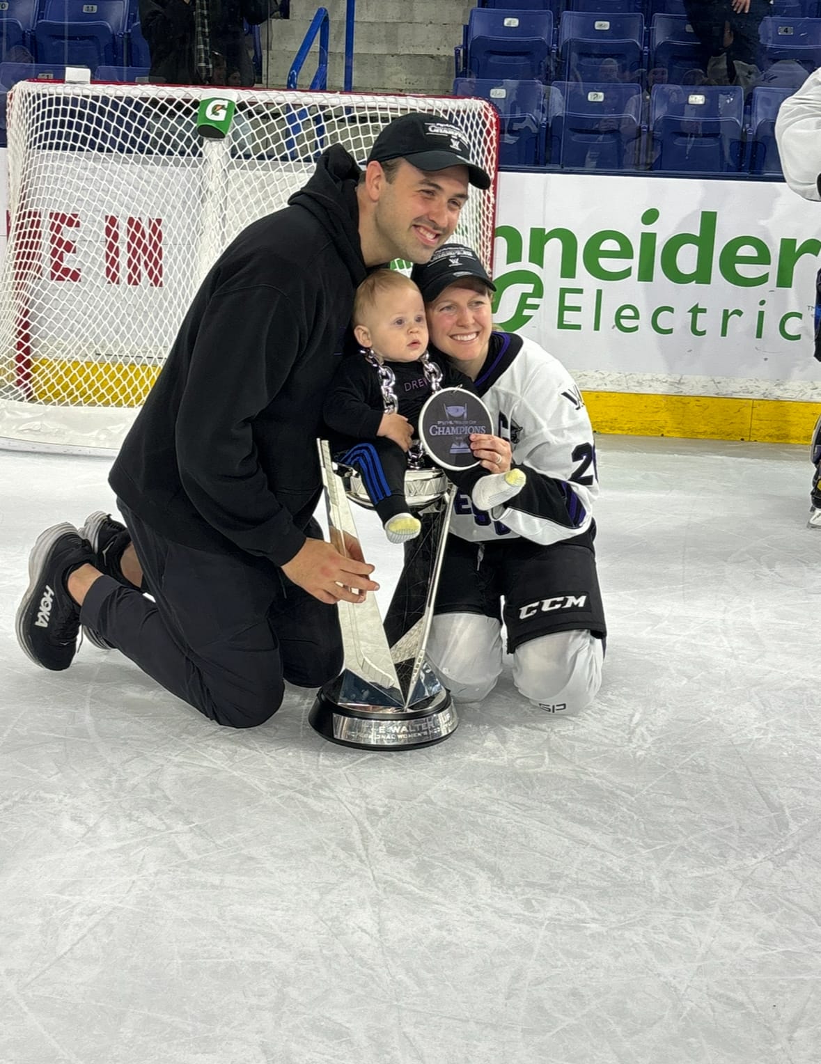 Coyne Schofield (right) and Schofield III (left) kneel on the ice while smiling towards photographers to the right of me. Their son is sitting in the Walter Cup in between them.
